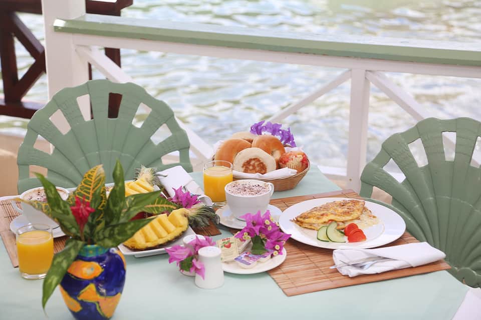 Beachfront breakfast table with tropical flowers, fresh pastries, juice, and ocean view beyond railing