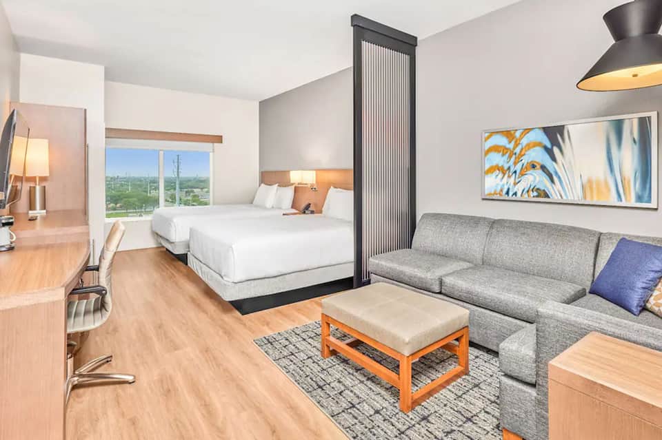 Modern hotel bedroom with two queen beds, sofa, city view window, and warm wood accents
