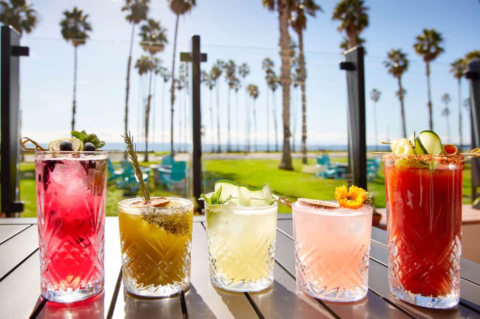 Five colorful cocktails on table with ocean, palm trees, and crosses in background