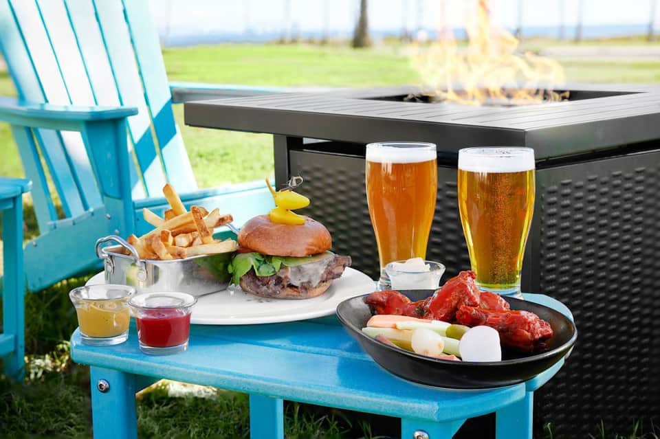Outdoor firepit table with burgers, appetizers, and beers beside a blue Adirondack chair