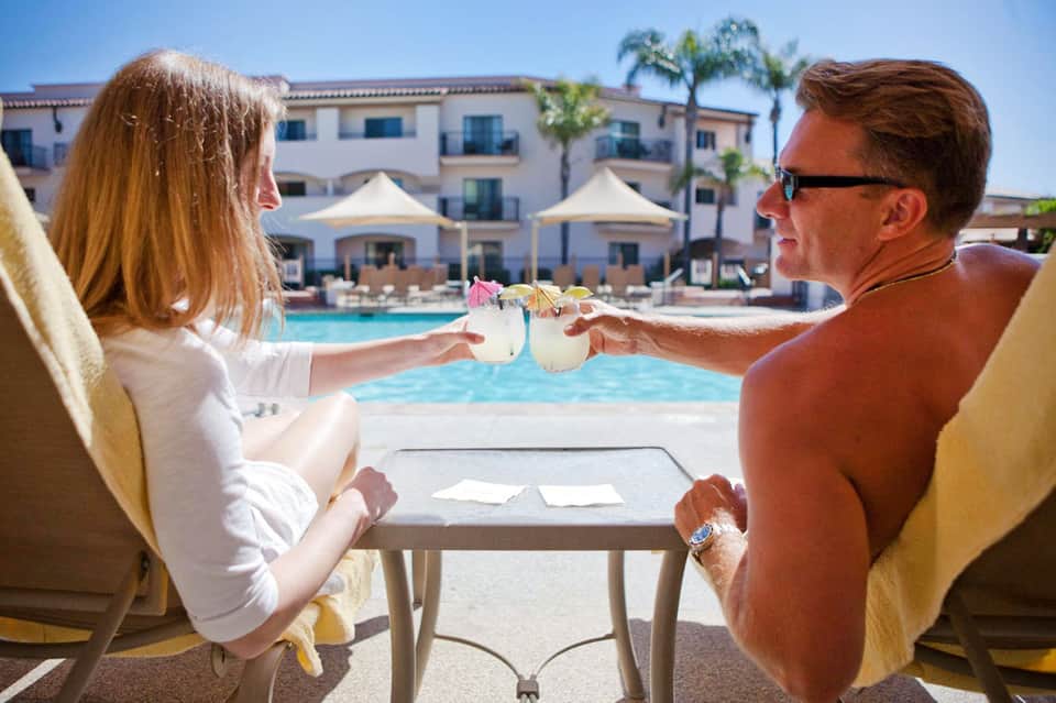 Couple enjoying tropical drinks by resort pool with Spanish-style building and palm trees backdrop