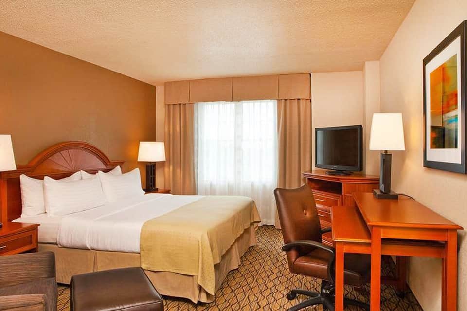 Hotel bedroom with queen bed, orange wood furnishings, desk area, and warm lighting