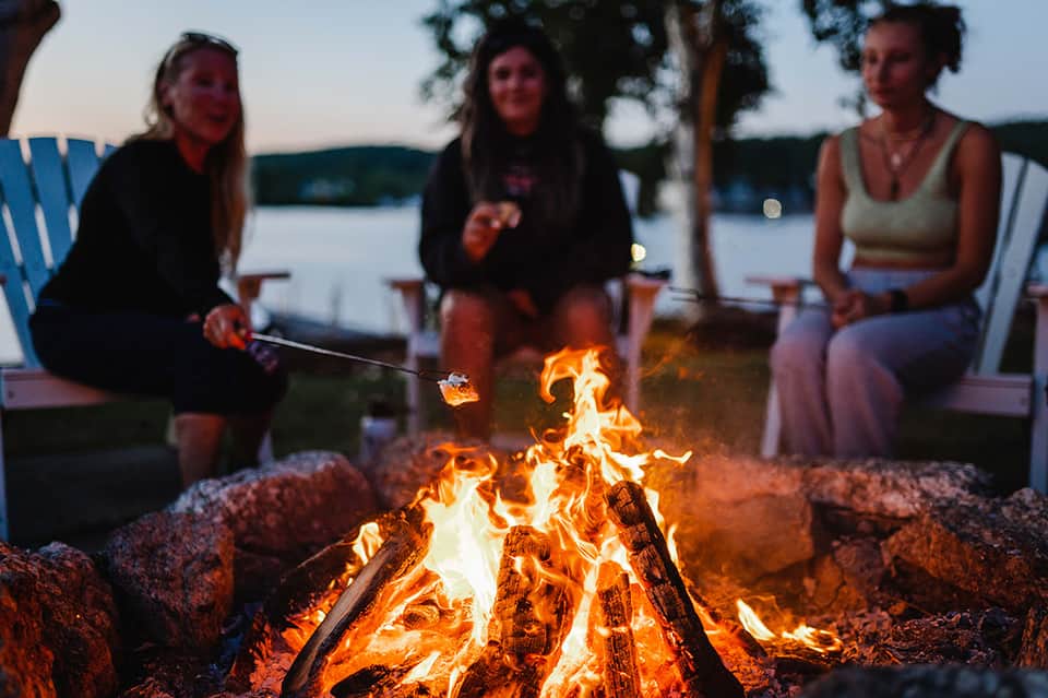Group of people roasting marshmallows around a campfire by a lakeside at dusk
