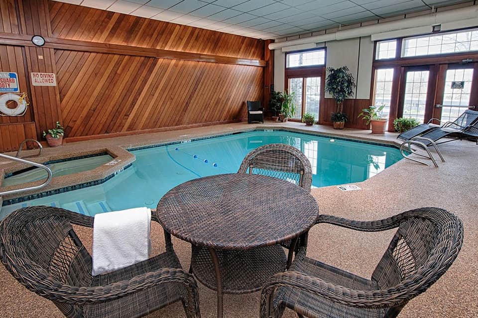Indoor pool facility with wooden vaulted ceiling, turquoise water, lounge seating, and potted plants.
