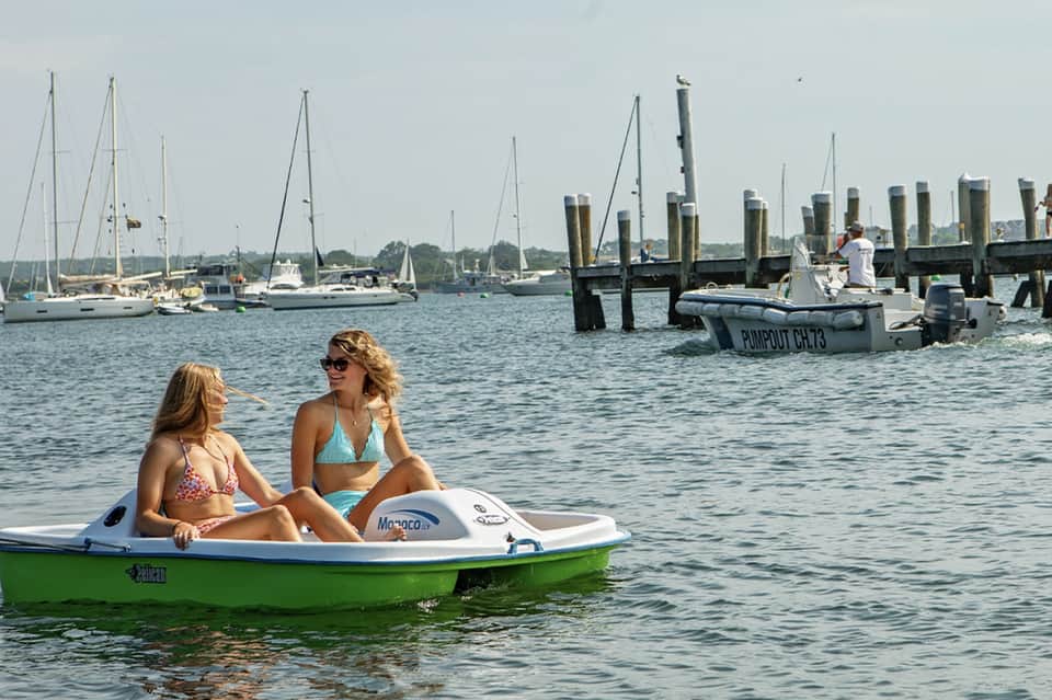 Two women on green motorboat with sailboats and dock pilings visible in calm harbor