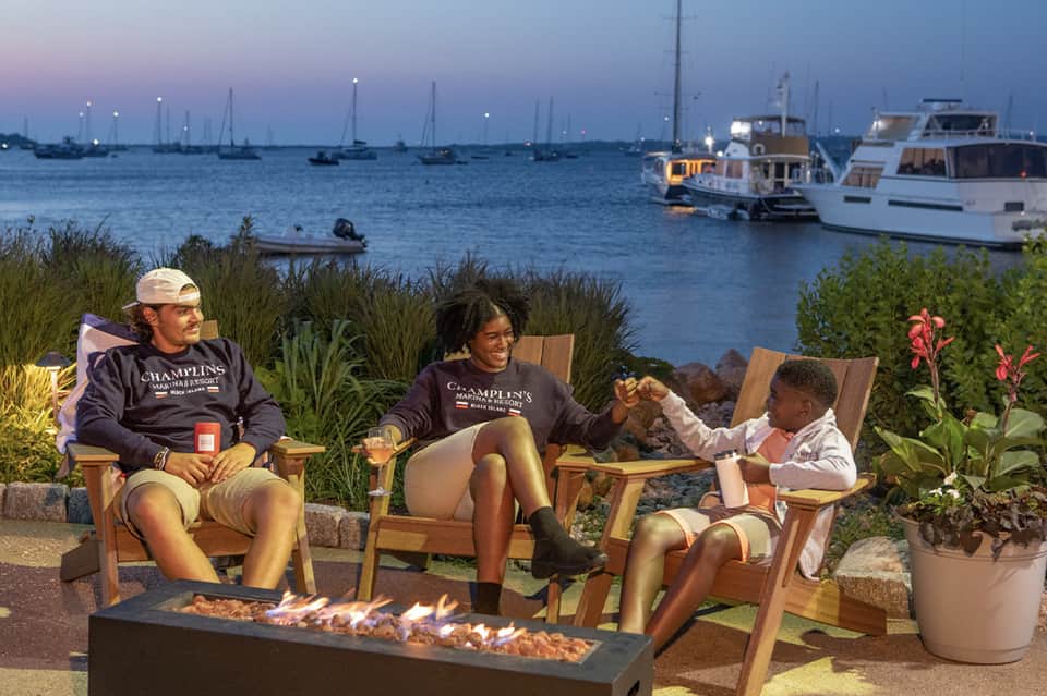 Family enjoying fire pit by the water at dusk with boats and marina visible