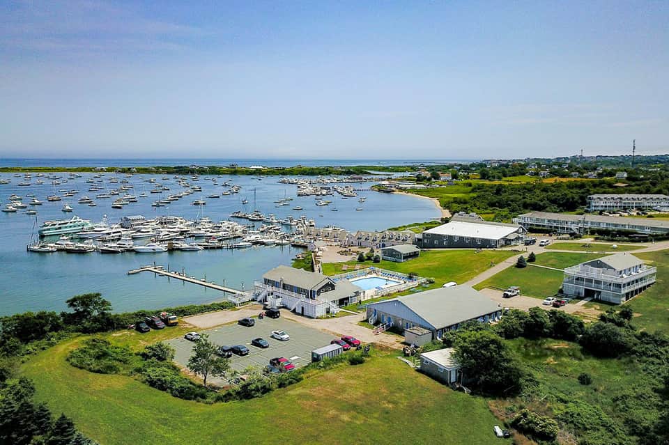 Aerial view of waterfront resort with moored boats, beach facilities, manicured lawns, and harbor views.