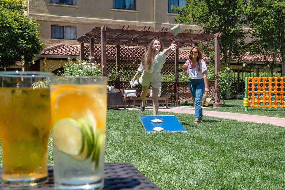 Women playing cornhole on lawn with drinks and pergola structure in background