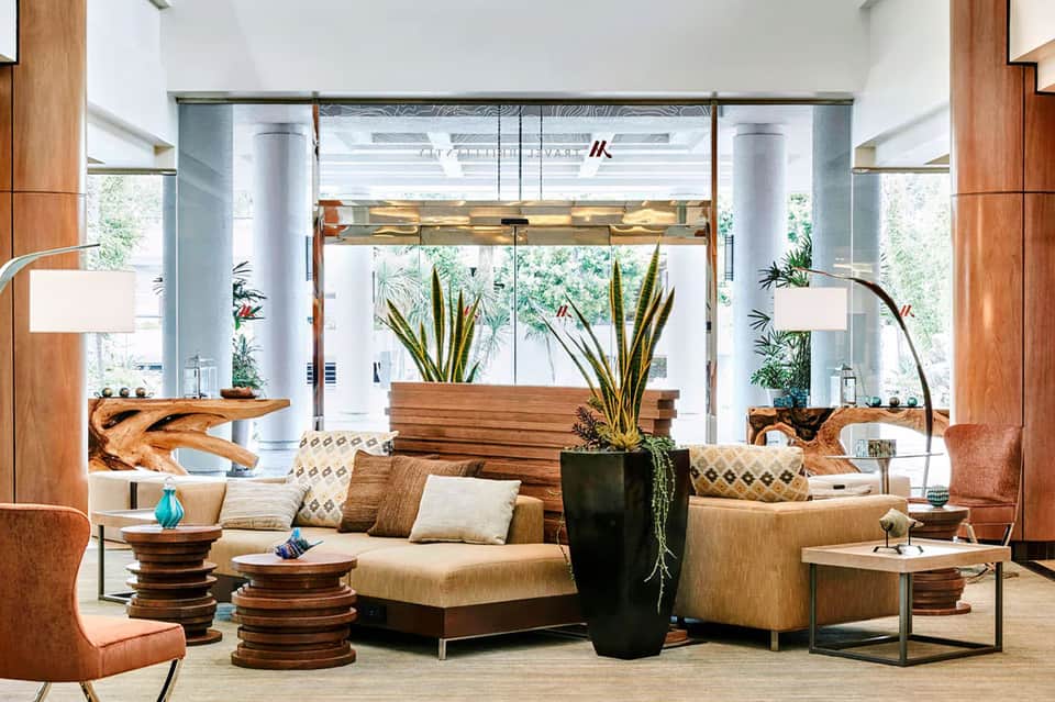 Modern hotel lobby with wooden accents, tan seating, tall windows, and indoor plants
