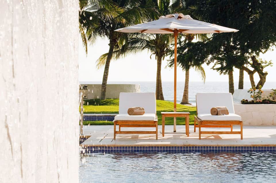 Poolside loungers with white cushions under wooden umbrella overlooking ocean and palm trees