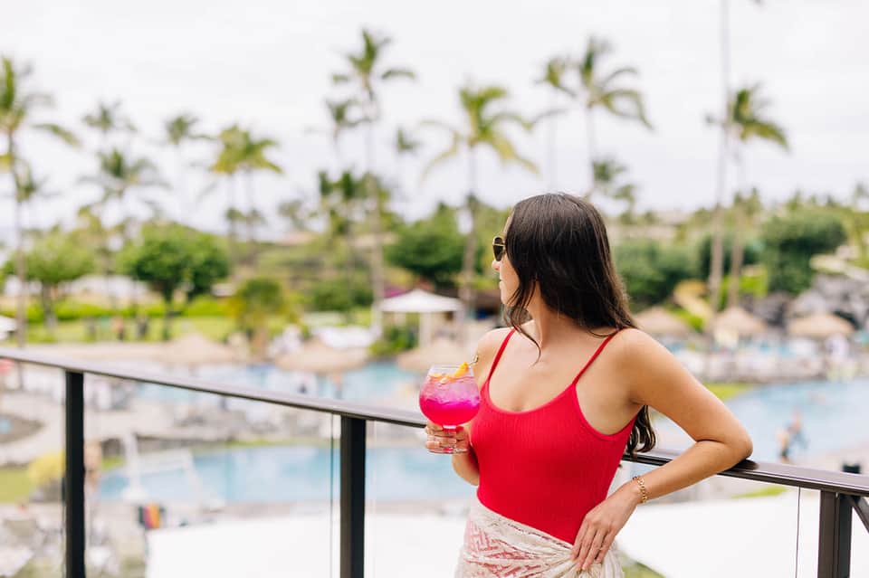 Woman on balcony holding pink cocktail overlooking resort pools and tropical landscaping below