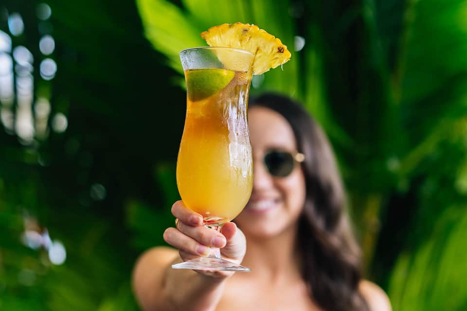 Woman in sunglasses holding tropical cocktail with pineapple garnish and green foliage