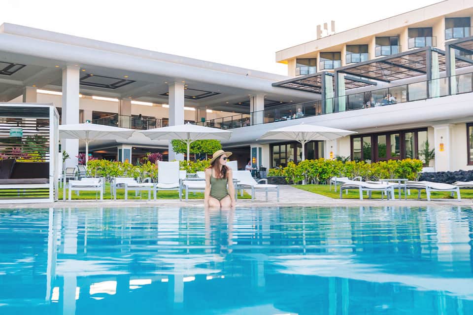 Woman in green swimsuit wading in resort swimming pool with white umbrellas and building