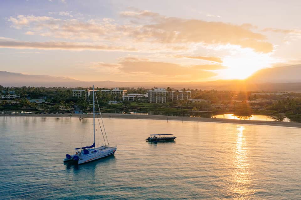 Sailboat at sunset with coastal town and mountains reflected in calm water