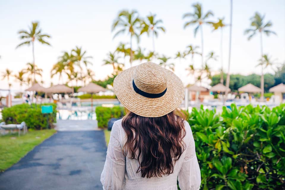 Woman in white shirt and straw hat overlooking palm-lined resort pool area
