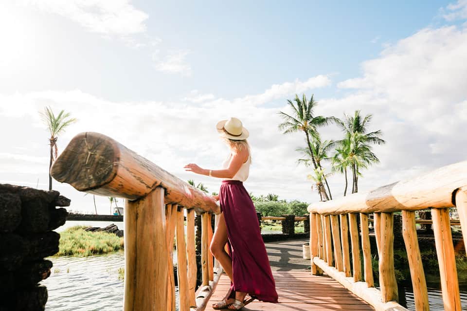 Woman in burgundy skirt and straw hat standing on wooden boardwalk with large wooden telescope sculpture overlooking water