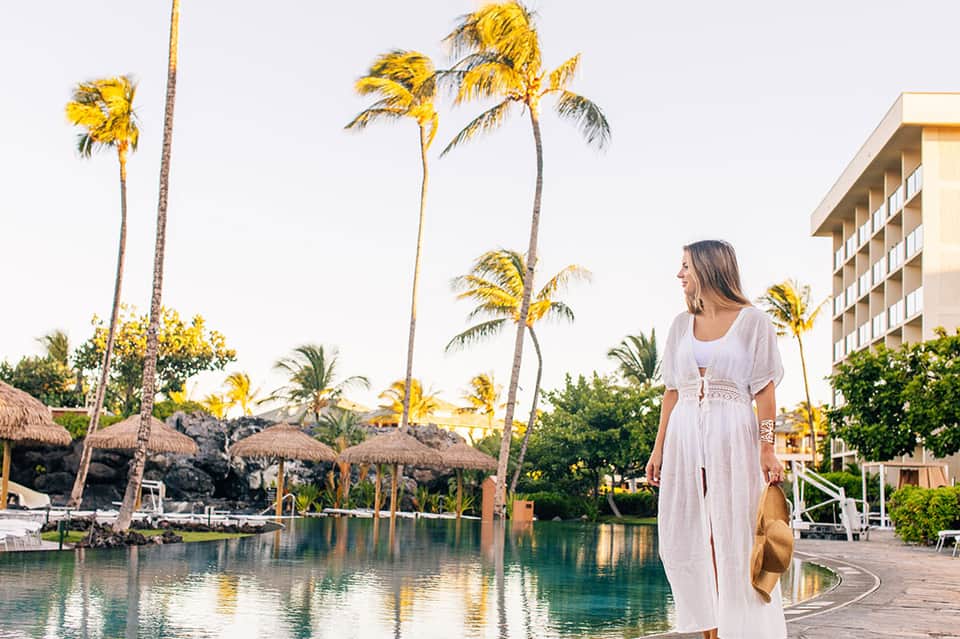 Woman in white dress at tropical resort pool with palm trees, thatched umbrellas, and hotel building background