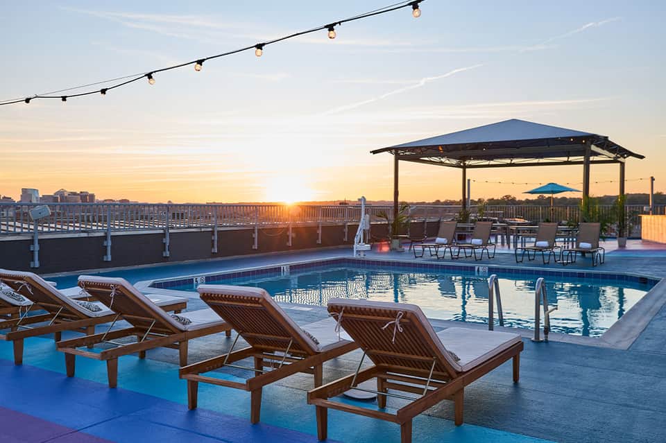 Rooftop pool at sunset with wooden loungers, gazebo, and city skyline silhouette