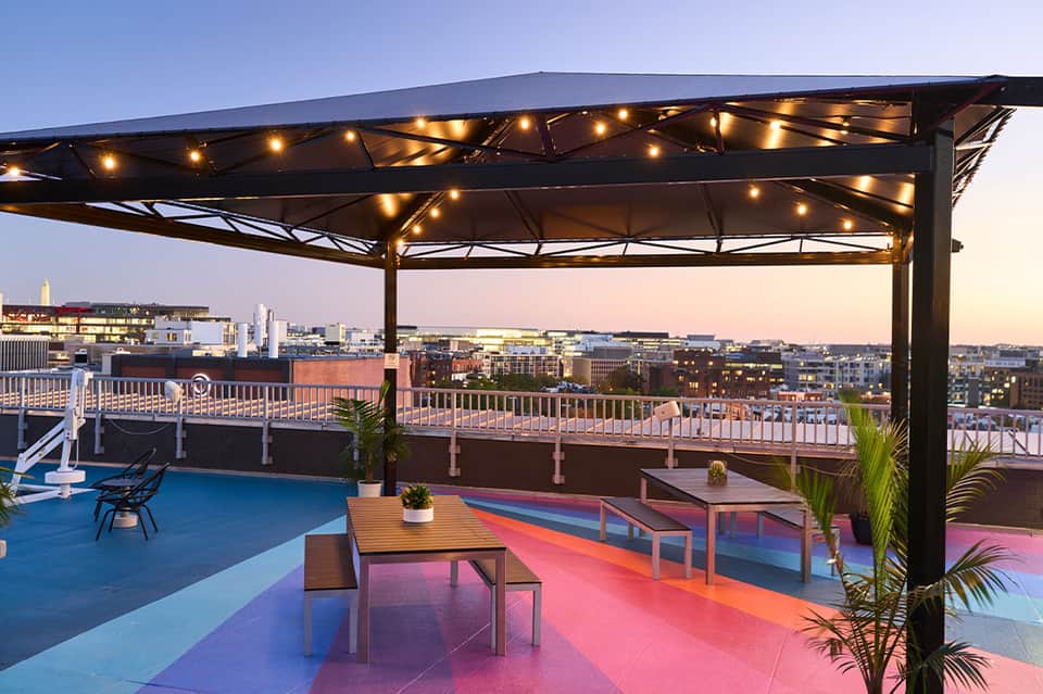 Rooftop lounge at dusk with colorful geometric floor, string lights, and city skyline views