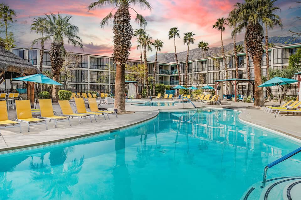 Expansive resort pool at sunset with yellow lounge chairs and palm-lined courtyard