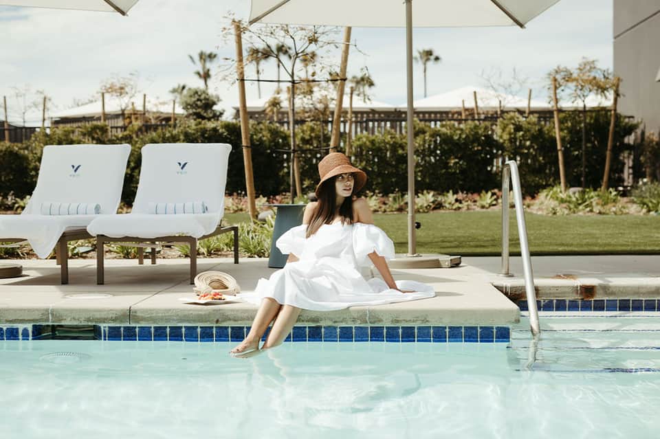 Woman relaxing by resort pool in white dress with palm trees and manicured lawn