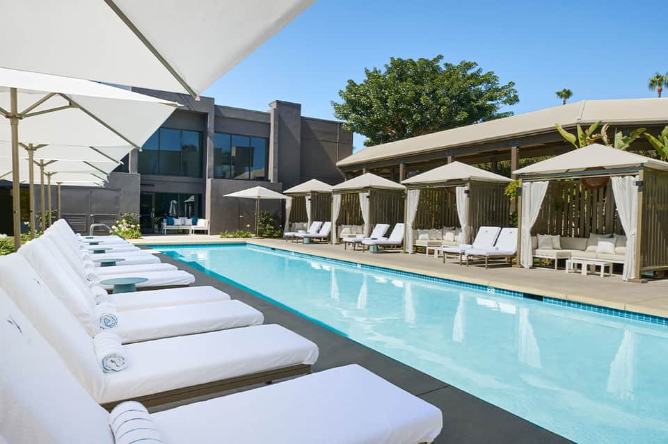 Resort pool lined with white lounge beds, cabanas with white curtains, and palm trees in bright sunshine