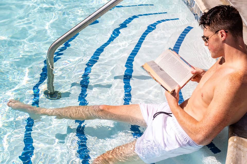 Man reading by swimming pool edge with blue lap lanes, relaxing in white swim trunks at resort pool