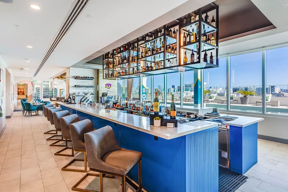 Modern rooftop bar with blue counter, full liquor display, and city skyline views