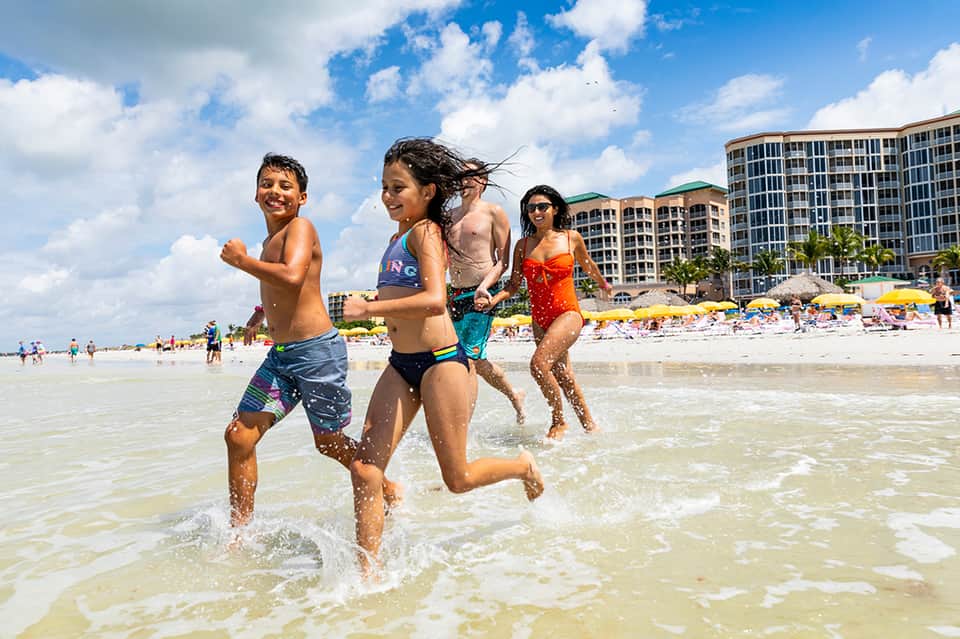 Children and adults running into shallow ocean water with beach and high-rise hotels in background