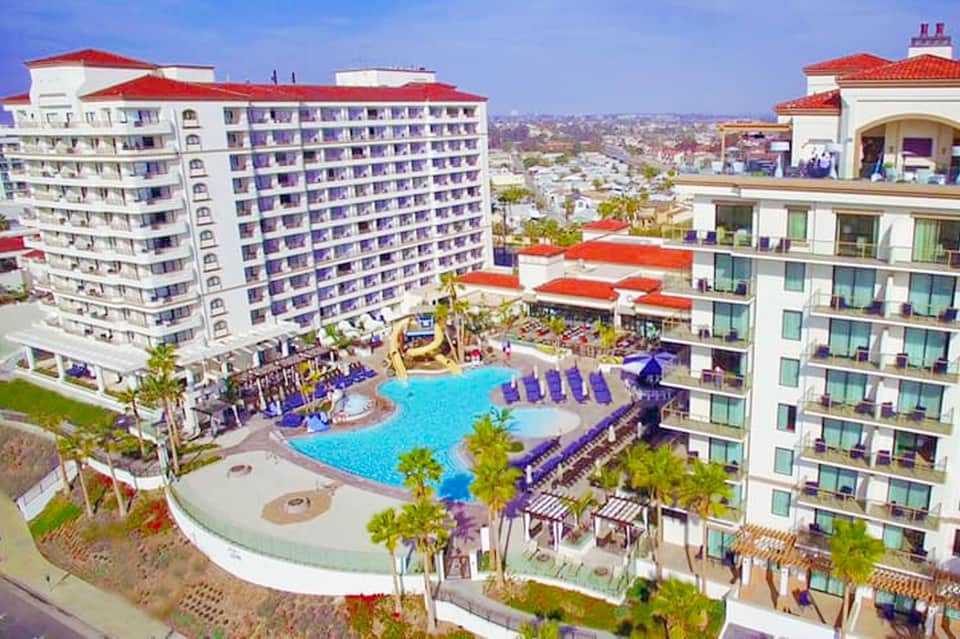 Aerial view of beachfront resort with white buildings, red roofs, and curved blue swimming pool surrounded by palm trees