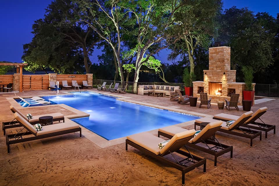 Illuminated resort pool at night with lounge chairs, fireplace, and mature trees