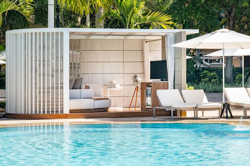 Modern poolside lounge with white cabana structure, furnishings, umbrellas, and crystal-blue pool water