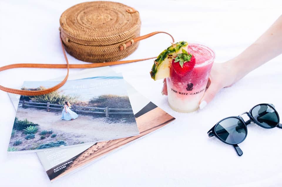 Hand holding pink cocktail with pineapple and strawberry garnish, sunglasses and woven bag nearby