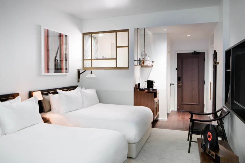 Modern hotel room with two white beds, wooden mirror, desk, and contemporary furnishings