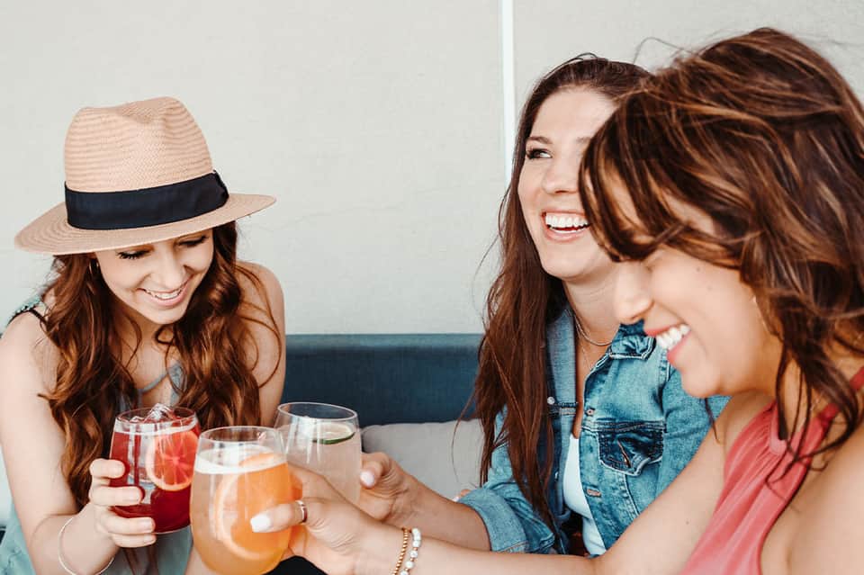 Three women with drinks laughing together indoors, one wearing straw hat