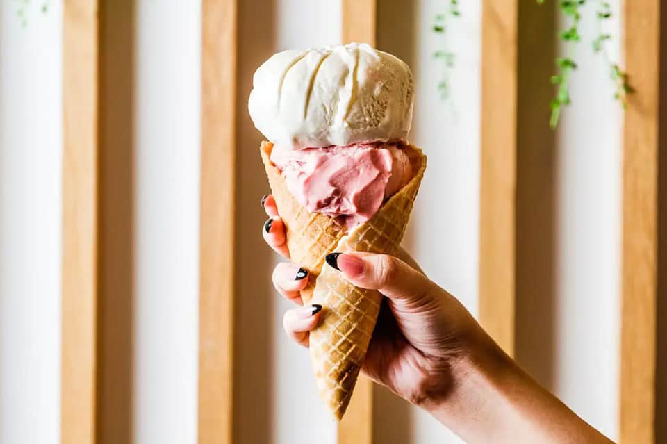 Hand holding three-scoop ice cream cone with pink and white flavors in sunlit setting