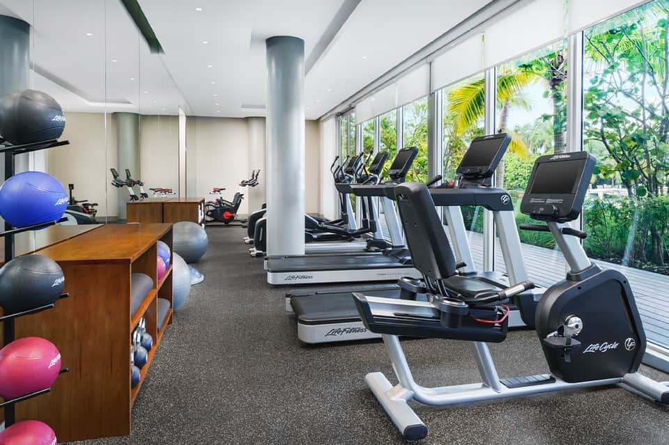 Modern fitness center with treadmills, exercise bikes, and floor-to-ceiling windows overlooking tropical landscaping