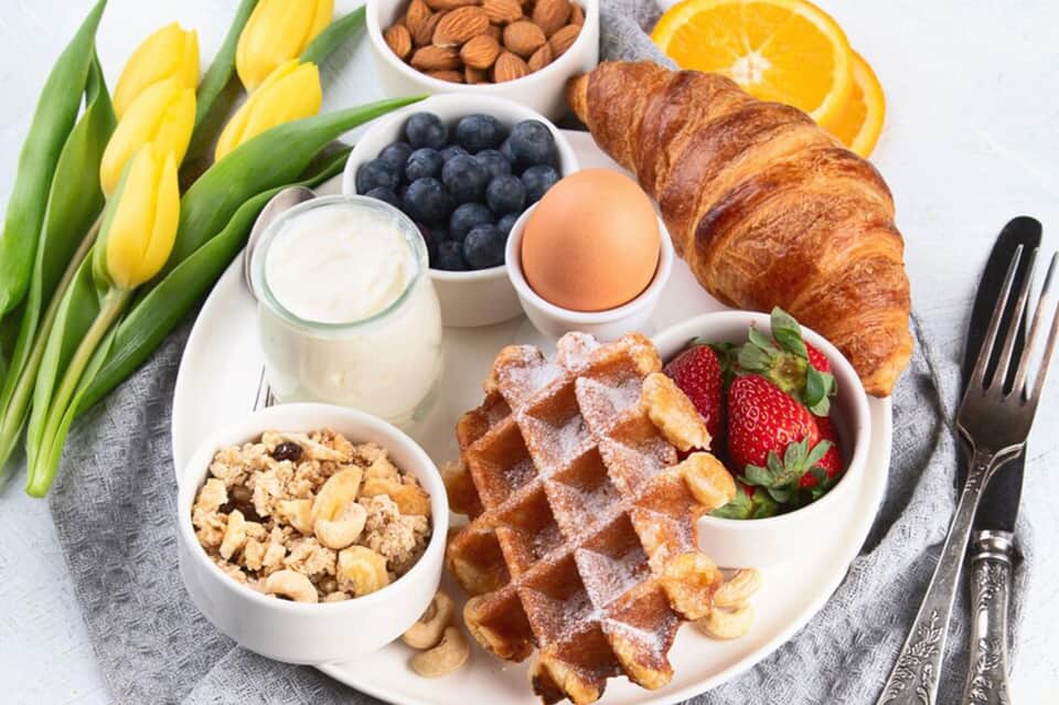 Breakfast spread with croissant, waffles, berries, yogurt, eggs, and fresh flowers on white plate