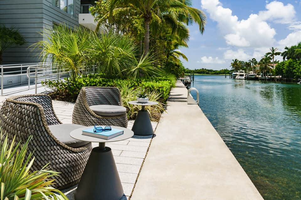 Waterfront patio with woven lounge chairs, black accent tables, tropical plants, and canal view on sunny day