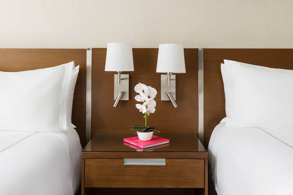 Modern hotel bedroom with two queen beds, wooden headboard, white bedding, and orchid centerpiece