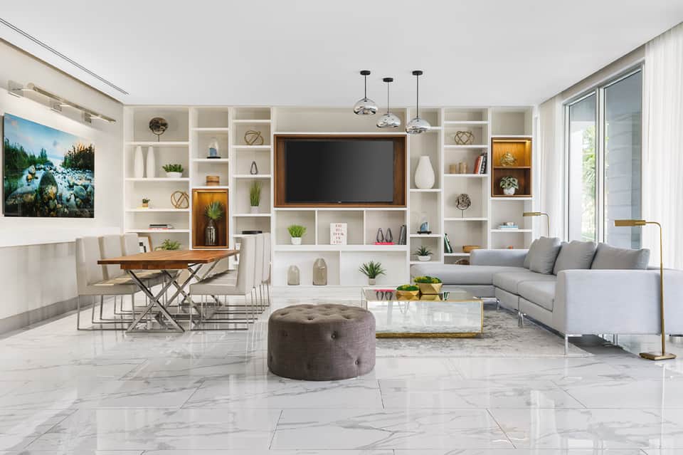 Bright modern living room with white built-in shelving, gray sofa, wooden desk, and marble flooring