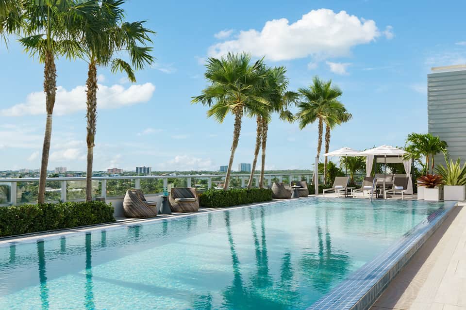Infinity-edge rooftop pool with palm trees and city skyline views with white loungers