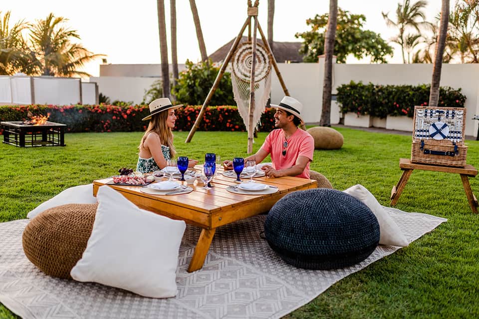 Couple dining on grass at wooden table with macramé swing and palm trees nearby