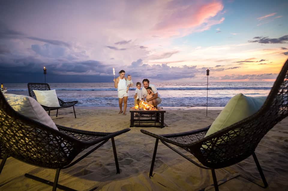 Family gathering around bonfire on beach at sunset with ocean and sky backdrop