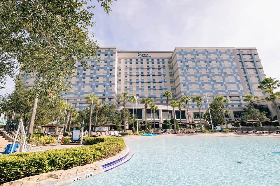 Large resort hotel with curved pool, palm landscaping, lounge chairs, and modern architecture by waterfront