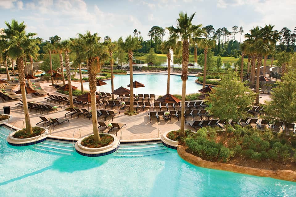 Large lagoon-style pool surrounded by palm trees, lounge chairs with dark umbrellas, and tropical resort grounds