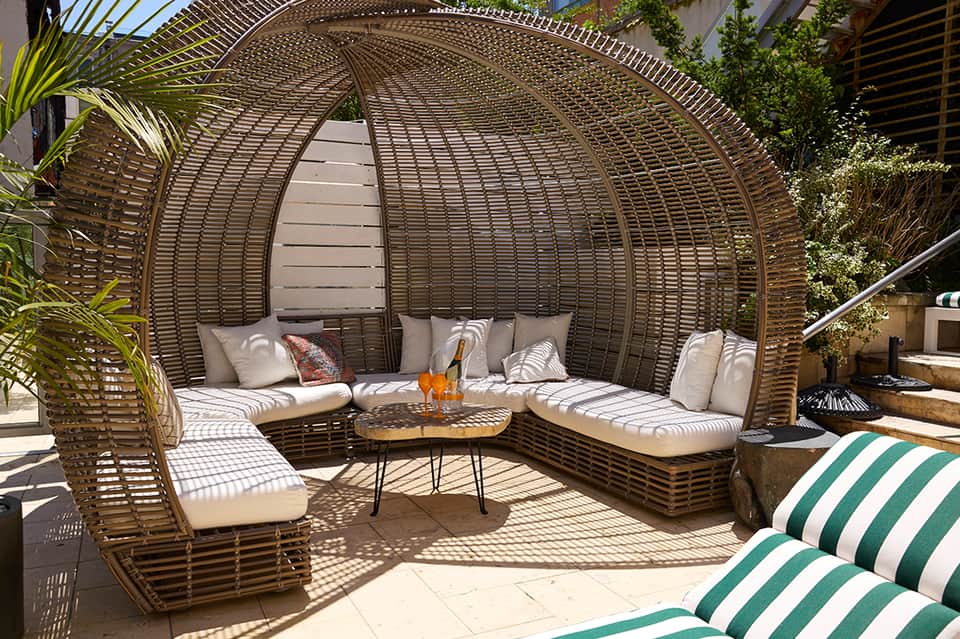 Woven rattan daybed pod with cushions, decorative pillows, wooden side table, and lounge chairs nearby