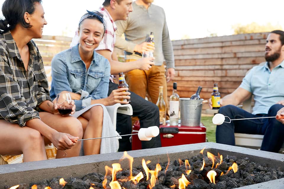 Friends socializing around outdoor fire pit with drinks and roasted marshmallows at dusk