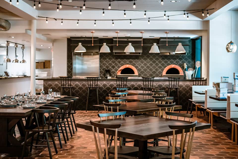 Modern restaurant interior with wood tables, pendant lighting, decorative tile wall, and two pizza ovens