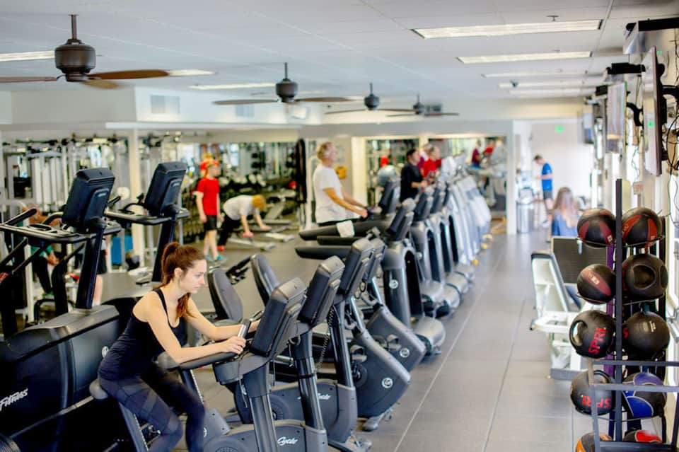Bright fitness center with numerous exercise bikes, equipment, and mirrored walls with people working out
